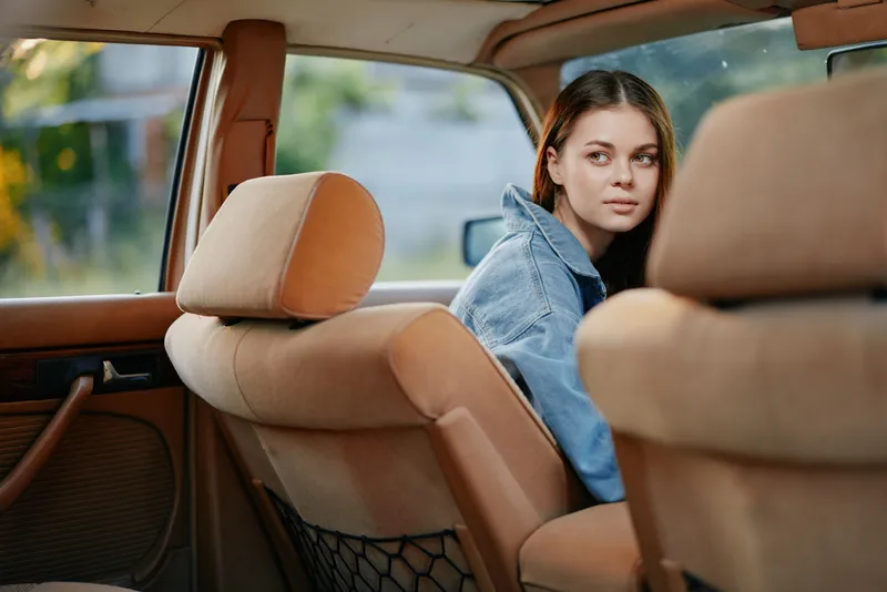 Young woman in denim jacket looking pensively out the car window, evoking a sense of nostalgia and contemplation against a warm background.