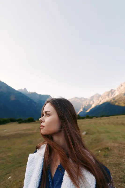 Woman portrait in profile with long hair in mountains meadow outdoors, nature landscape and travel scene at golden hour. Young female model looking thoughtful in open field.