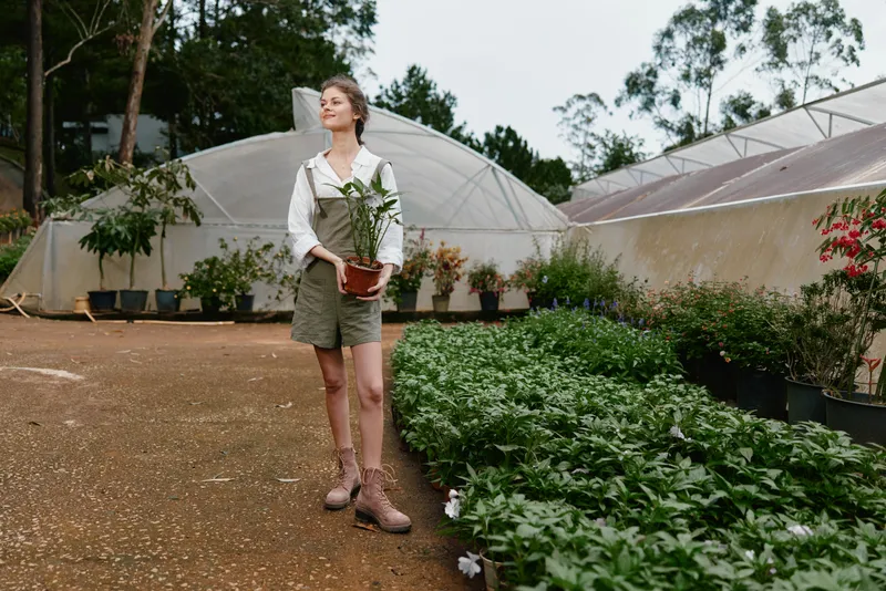 Young woman with brown hair, wearing a casual white shirt and green shorts, joyfully carrying a potted plant in a greenhouse setting, showcasing a connection to nature