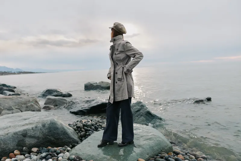Woman in coat and hat stands on rocky shore looking at sea. Overcast sky and calm water create serene outdoor scene with natural coastal landscape and peaceful mood.