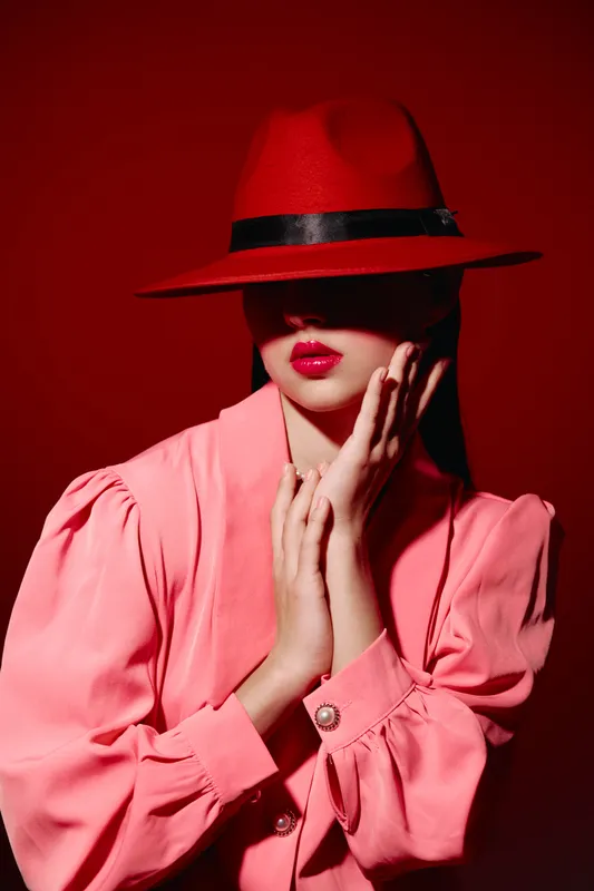 Young woman in red hat and bright pink blouse poses with her hands near face on red background. Creative beauty fashion concept focusing on bold colors and stylish expression.