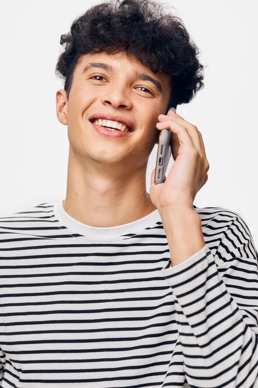smiling young man talking on smartphone wearing striped shirt with curly hair, bright background, casual and positive communication concept