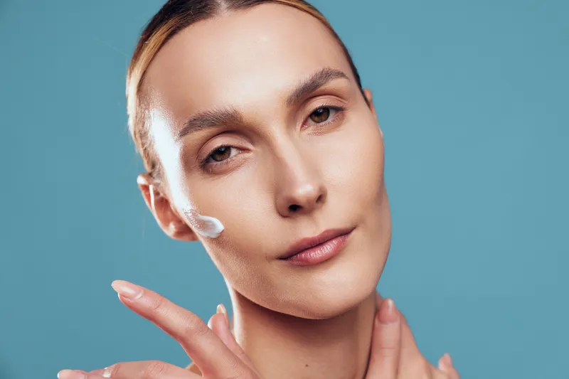 A close up of a woman with clear skin applying moisturizer on her cheek with a finger against a solid blue background. Concept for skincare, beauty, and self care routine.