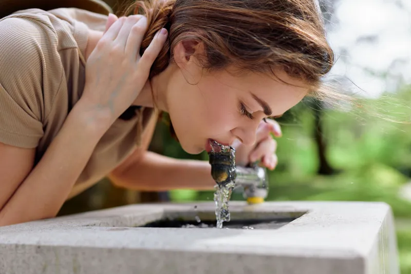 Woman drinking water from outdoor fountain in park, portrait closeup of young adult leaning over tap to sip, summer sunlight, casual clothing and natural hydration moment.