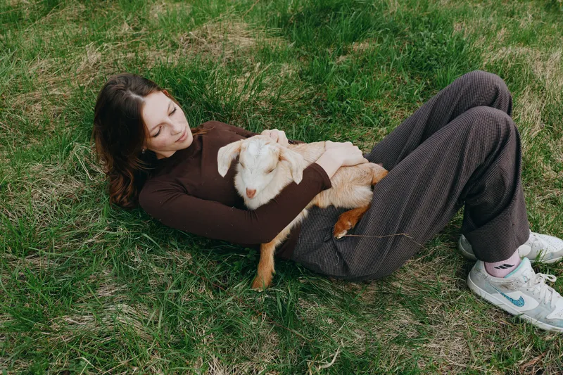 Young woman with brunette hair lies on grass holding a baby goat in her arms enjoying a peaceful moment outdoors in casual clothes and white sneakers