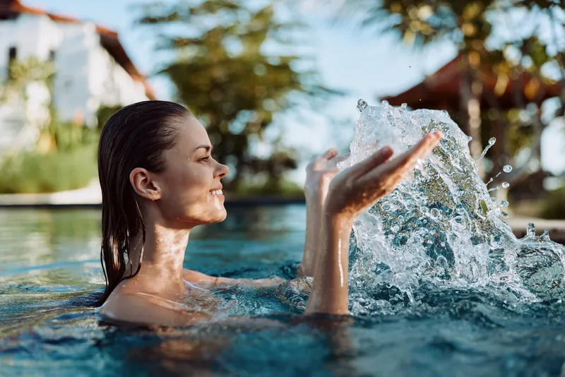Smiling woman in a pool splashes water with joyful motion, outside under bright sunlight, capturing a relaxed lifestyle moment. Clear blue water, warm colors, and a lively mood convey wellness,