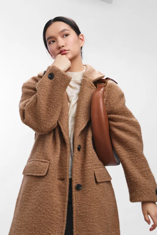 asian woman wearing a brown coat stands against a white studio backdrop, radiating confidence and poise as she surveys the surroundings, presenting a polished and contemporary fashion moment.