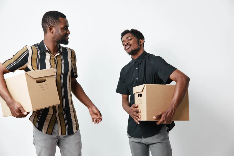 Two African American men with dark skin carrying cardboard boxes, smiling and interacting in casual clothes against white background for moving or delivery.