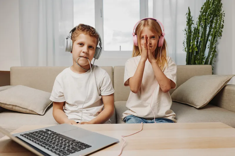 Two children with headphones sit on a couch in a bright living room, one boy looking serious, one girl covering her face with hands. Concept of childhood and technology.