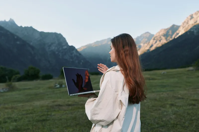 A focused outdoor professional stands with a laptop in a mountain valley. She wears light outerwear, gazing ahead with confidence, creating a calm, productive, and inspirational work scene.