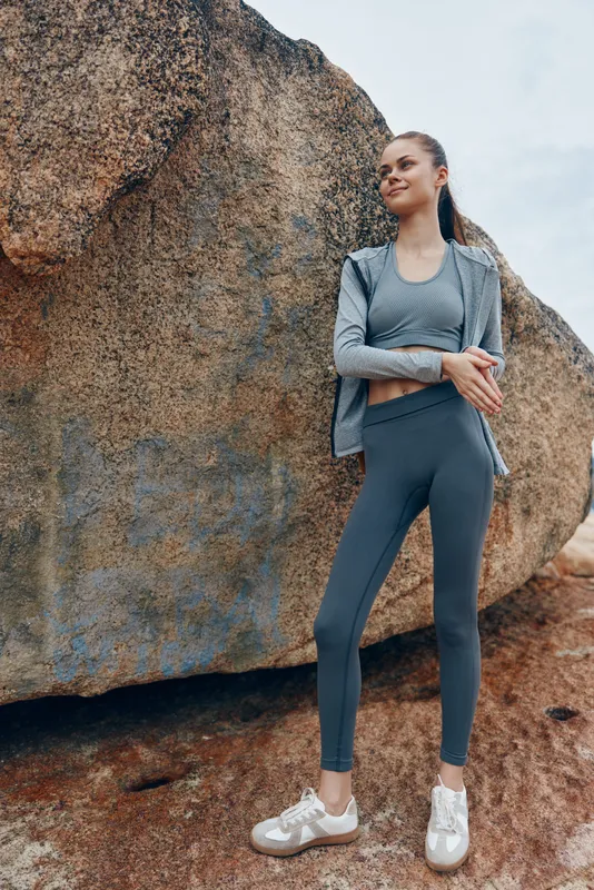 Stylish female model in activewear poses confidently against a large rock, showcasing a serene expression and vibrant gray outfit in a natural outdoor setting