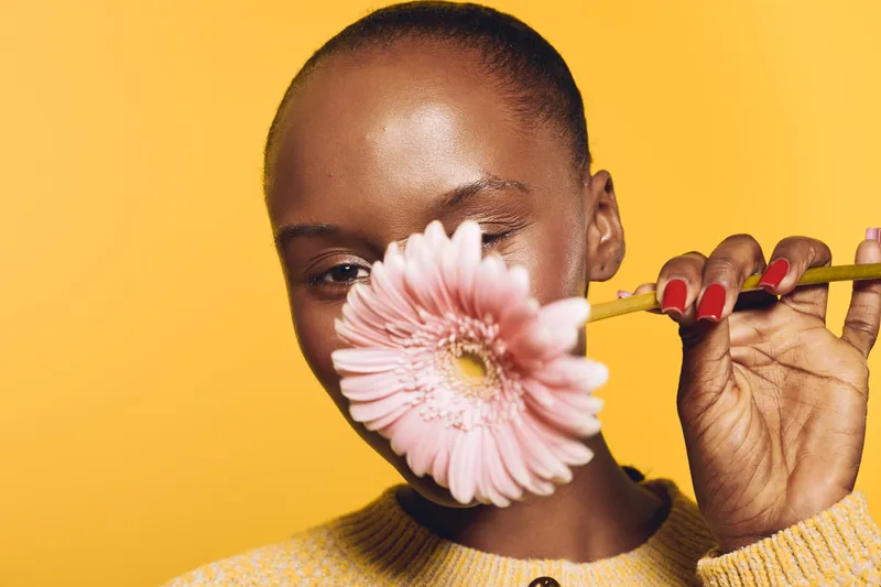 Close-up of a smiling dark skinned African woman holding a pink flower in front of her face against yellow background. Concept for beauty, nature, happiness, and floral portrait.