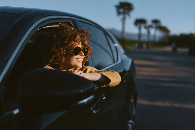 Smiling woman with curly hair leaning from a car window, wearing sunglasses. Sunset light paints an open road with palm trees, emphasizing a relaxed lifestyle and carefree moment.