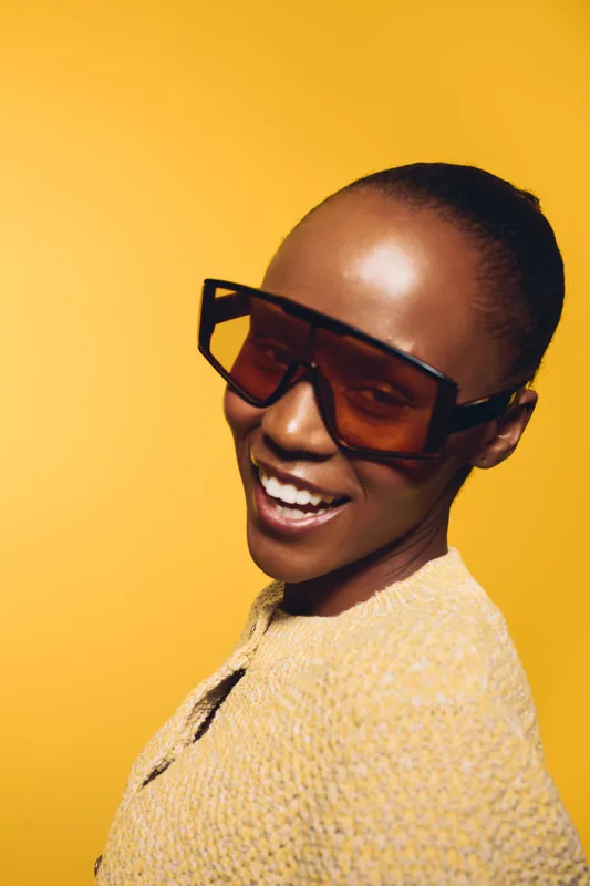 Smiling African American woman with dark skin wearing oversized sunglasses and a yellow sweater poses against a bright yellow background. Concept of fashion, confidence, and style.