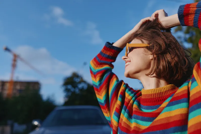 Woman in colorful striped sweater smiling with sunglasses, young adult with short hair looks up against urban blue sky, hands in hair, casual travel lifestyle portrait.