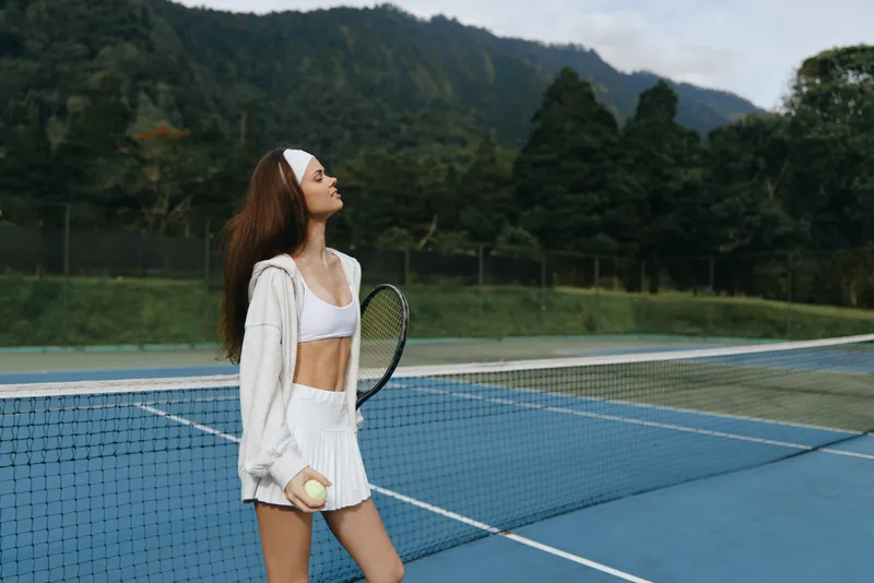 Young female tennis player in sportswear with a racket on a tennis court, exuding confidence and determination amidst a scenic mountain backdrop