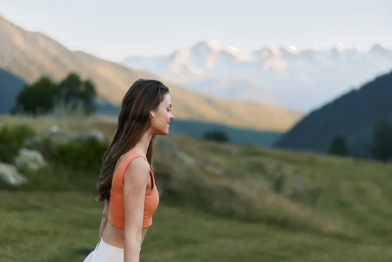 A cheerful woman stands in a scenic meadow near distant mountains. She wears an orange top and smiles softly, creating a calm, uplifting outdoor mood and a sense of freedom.