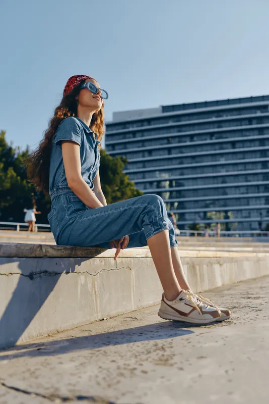 Lifestyle woman in modern boho-western denim street style sitting outdoors with sunglasses and a red bandana, captured in warm film color tones for a casual urban vibe.