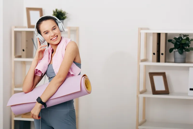 Athletic woman in workout wear with a yoga mat and towel, wearing headphones, smiling after a session in a bright room with minimalist shelves.