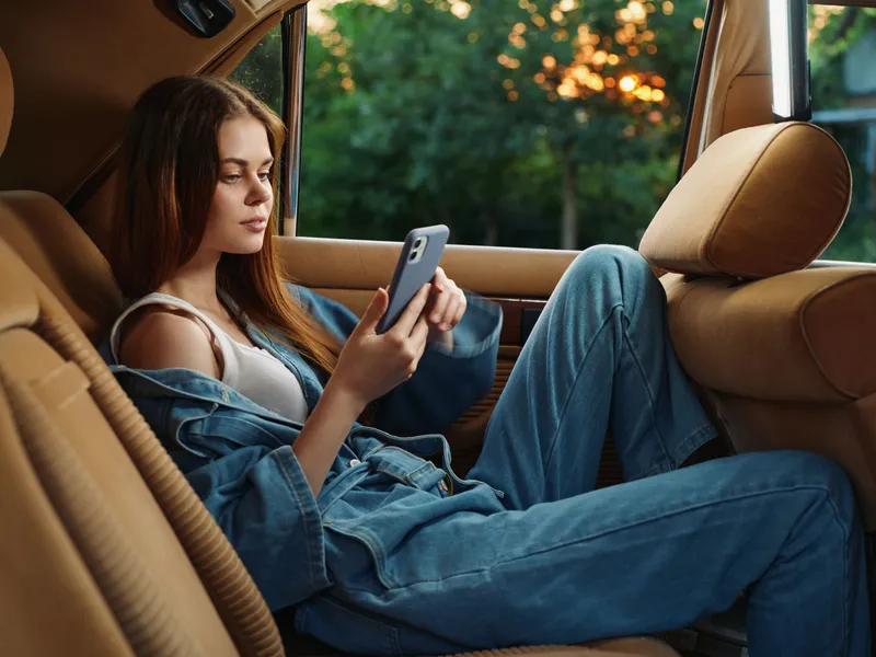 Young woman in denim outfit relaxing in a car during sunset, using her smartphone, showcasing a calm and carefree emotion, with a beautiful natural background.