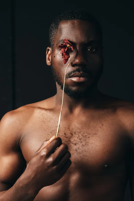 Creative conceptual portrait of a shirtless black man holding a dried flower over one eye in a dark studio. Artistic visual style with strong emotional expression and minimalistic background.