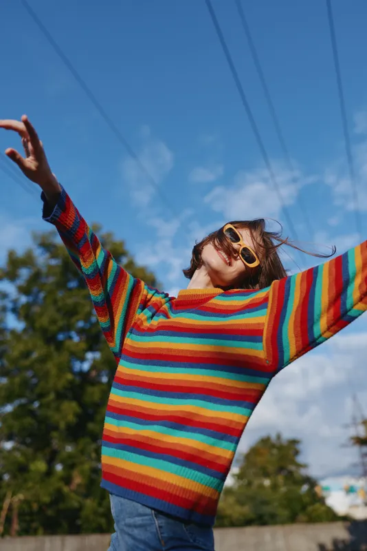 Woman young adult in her 20s wearing rainbow striped sweater and yellow sunglasses, smiling with arms raised outdoors under a sunny blue sky in a suburban street, joyful casual outfit lifestyle