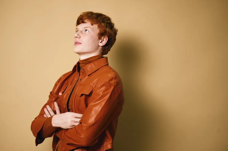 Young man with red hair wearing a brown leather jacket stands with crossed arms looking upwards on beige background. Casual style, confident pose and thoughtful expression.