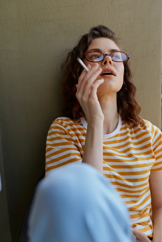 Casual woman portrait, relaxed mood as she lounges by a neutral wall, striped shirt, sunglasses, cigarette in hand, soft light, urban vibe, home lifestyle