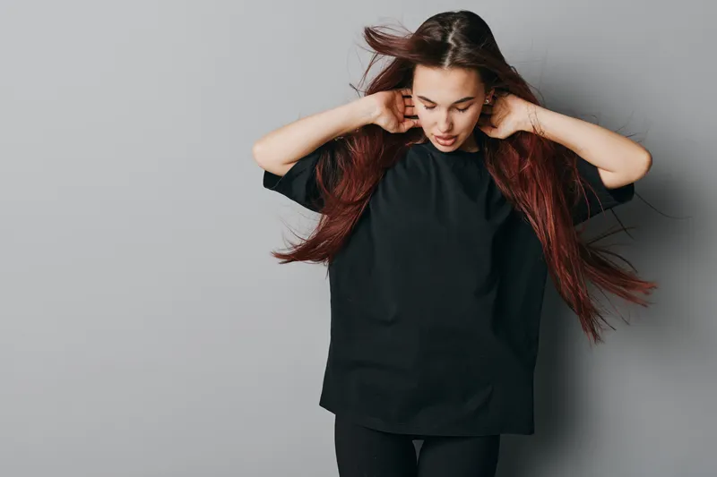 Young woman with long red hair wearing black oversized t shirt and pants standing against gray background looking down with hands near neck in casual style portrait.