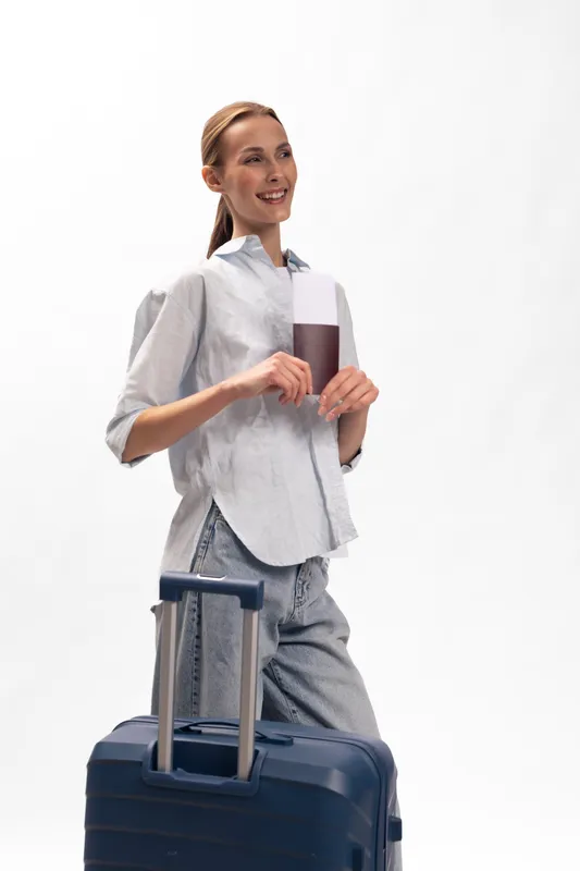Smiling woman with suitcase holds passport and boarding pass, ready for travel. Casual outfit and ponytail hairstyle highlight her excitement and preparation for a trip. Concept of tourism and