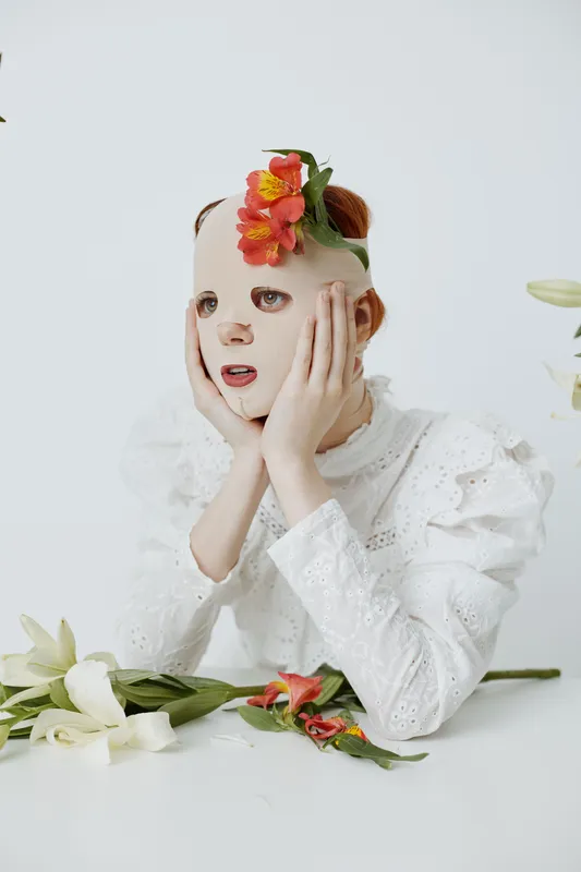 A model wearing a white dress and a floral mask, expressing emotion in a minimalist setting, symbolizing identity and beauty in a seasonal concept
