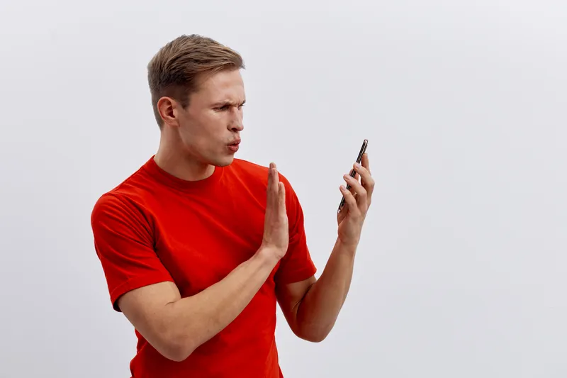 Man looking at mobile phone with surprised expression, wearing casual red t-shirt, isolated on plain white background, studio portrait, people lifestyle concept, communication, technology, astonishmen