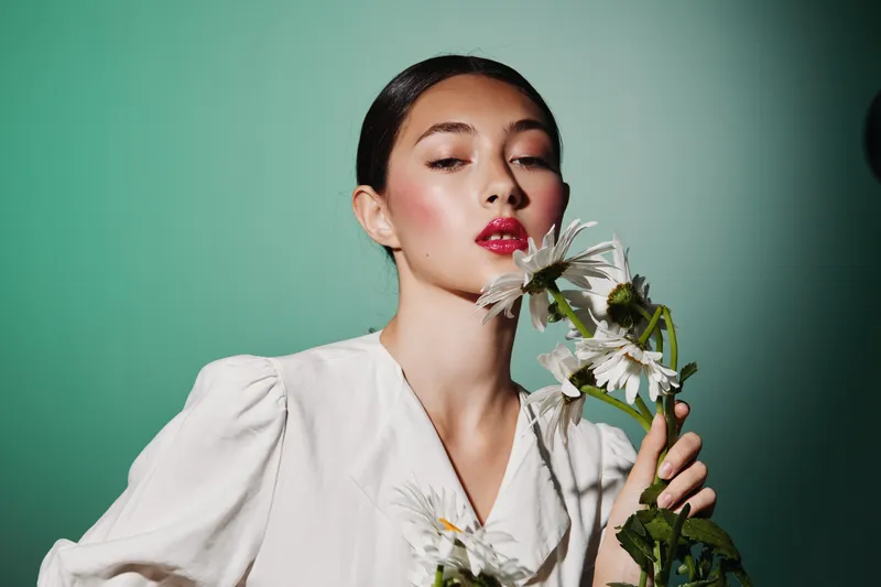Stylish young woman with glossy makeup and vibrant red lips poses holding white flowers. Soft green background contrasts elegant white blouse. Concept for fashion and beauty creative.