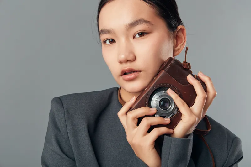 Young woman holding vintage camera with leather case, wearing gray jacket and posing with neutral expression on gray background in studio photography concept with focus on portrait.