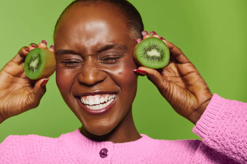 Happy dark skinned African woman with shaved head holds kiwi halves near ears, smiling with closed eyes. Bright green background and pink sweater create vibrant fresh concept of health and joy.