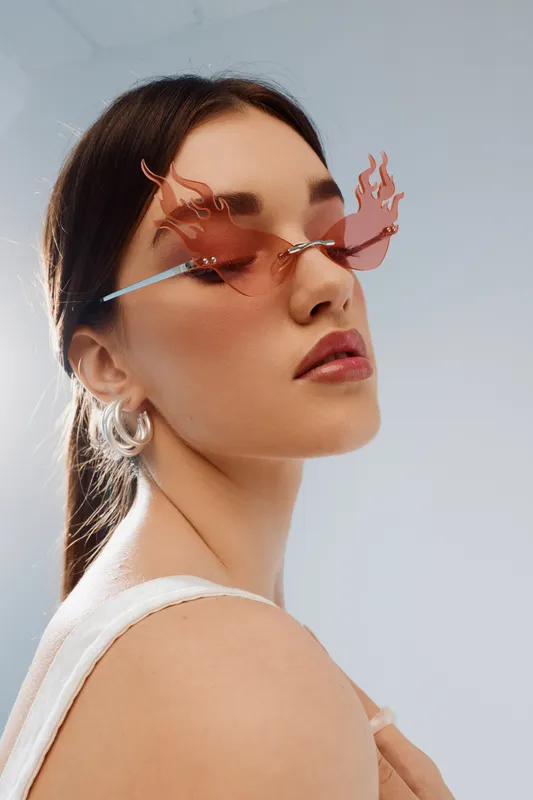 Young woman with long hair wears stylish flame-shaped sunglasses and white top. Close-up portrait on light background. Concept of woman beauty fashion and modern accessories.