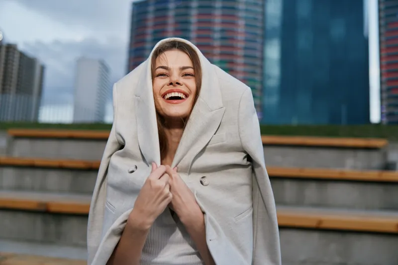 Happy businesswoman in a light-colored coat, smiling against an urban backdrop, conveying confidence and positivity in a dynamic business environment.