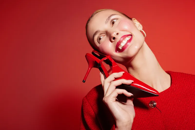 Smiling woman in red sweater holds red high heel shoe near face, looking up with bright lipstick on red background. Concept of fashion, beauty, and joyful mood.