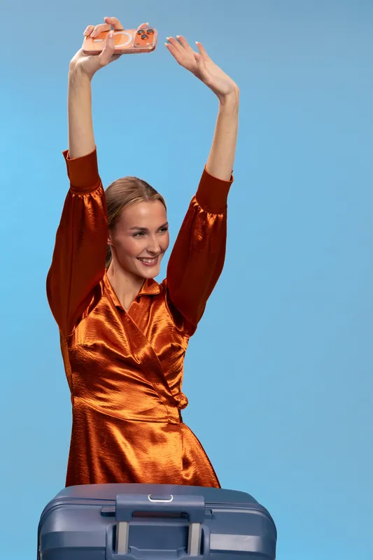 Happy woman in orange satin dress holds small bottle above head smiling, blue background. Concept for travel, vacation, packing, holiday, tourism, and joyful moments.