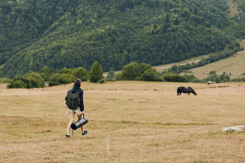 A lone hiker with a backpack walks across a wide meadow toward a grazing horse in a mountainous landscape, evoking adventure and solitude in a rustic outdoor setting under a clear sky.