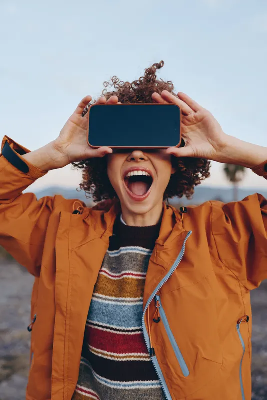 Woman outdoors laughing with smartphone held over eyes, sunny sky background, casual jacket and striped sweater, curly hair visible, joyful lifestyle moment in bright light