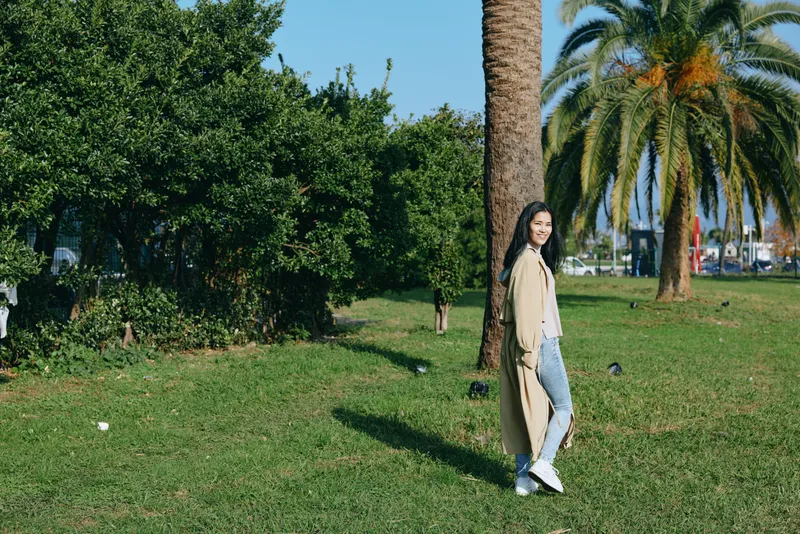 Woman in light coat and jeans strolling in a sunny park with palm trees, green grass and shade, conveying casual outdoor style, leisure and natural confidence