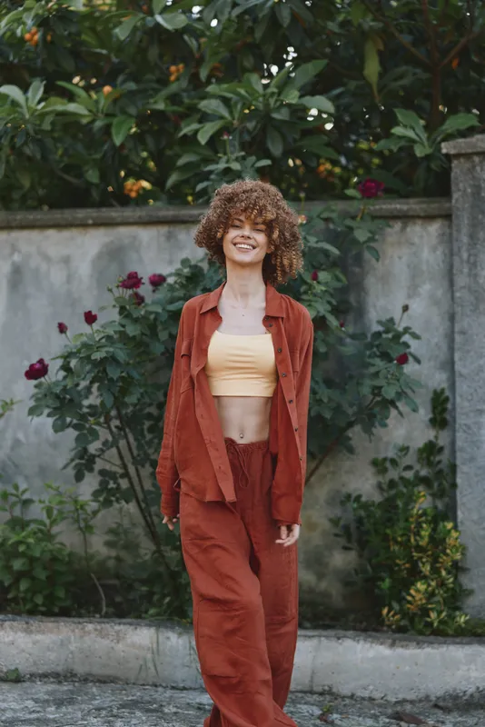 Casual confident woman with curly hair walks outdoors in an orange suit and crop top, posing near a garden wall, a candid lifestyle fashion moment under sunlight