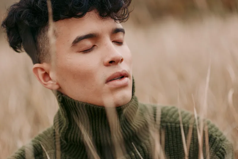 Close-up portrait of a young man with closed eyes in tall grass, soft natural light and sincere expression that highlights authenticity and conveys quiet credibility in a candid outdoor moment