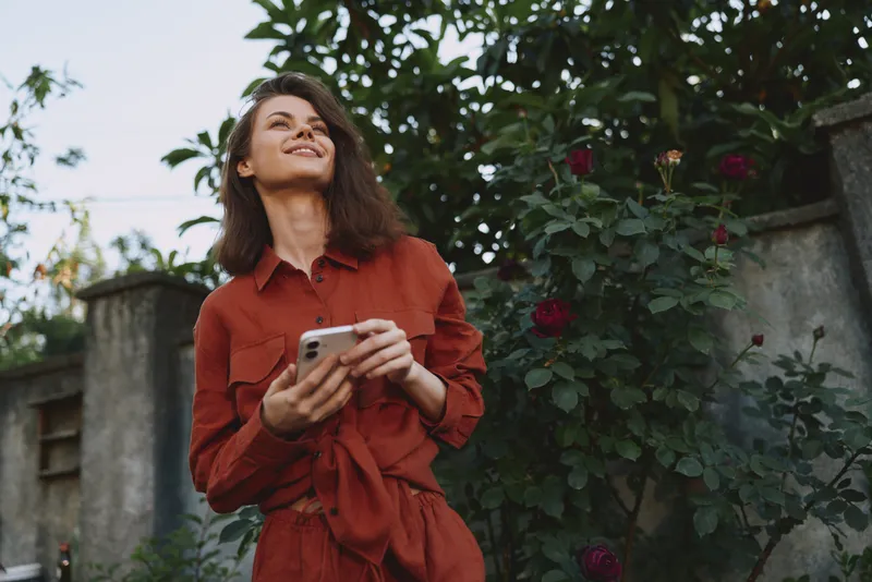 Woman lifestyle outdoor portrait in a red dress holds a smartphone, smiling and looking up amid a garden with roses and greenery, joyful candid moment