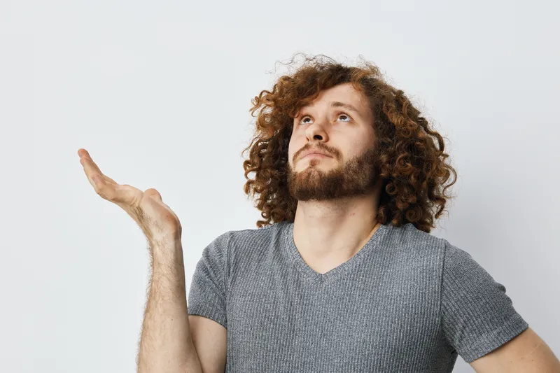 Thoughtful young man with curly hair contemplating in a gray shirt against a neutral background, expressing curiosity and introspection