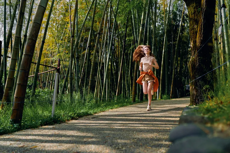 Woman running on a sunlit bamboo path in a green forest, trail motion captured with flowing hair and active fitness lifestyle, outdoor exercise, freedom and joyful movement.