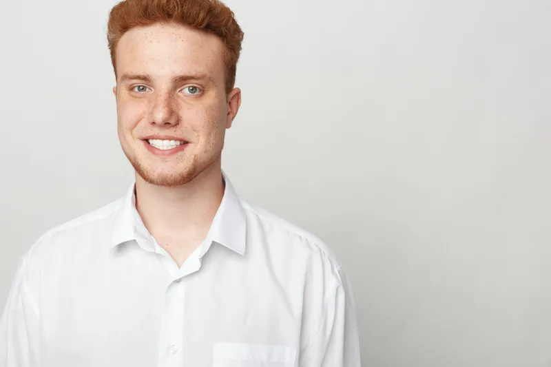 Smiling young man with red hair and freckles wearing a white shirt stands against a plain light background. Portrait of confident male model with casual style and natural expression.