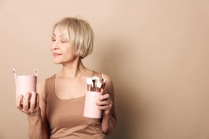 Mature woman with short blonde hair holds pink containers with makeup brushes, wearing beige tank top. Concept of beauty routine, self-care, and elegant skincare for older adults.