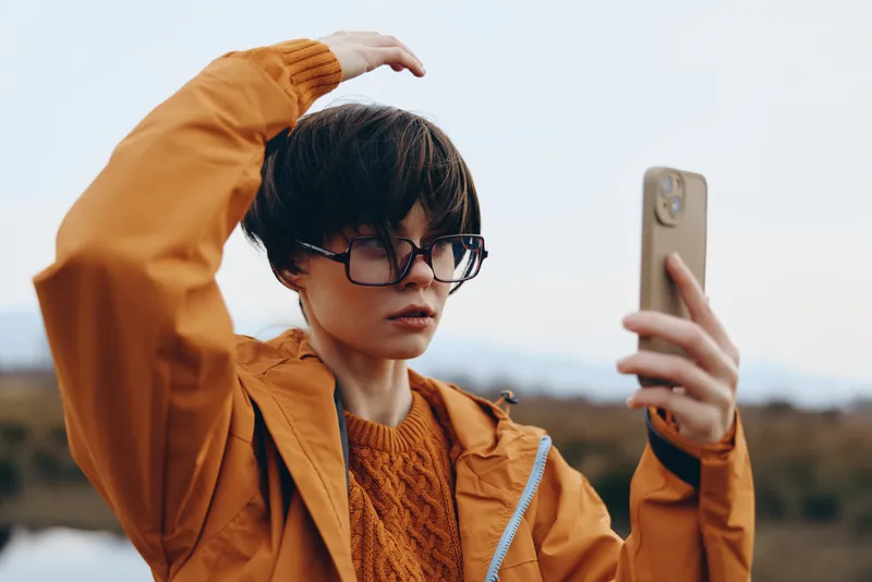 Woman lifestyle outdoors, taking a selfie with phone, adjusting hair with one hand, wearing glasses and an orange jacket, calm expression, soft daylight, blurred background